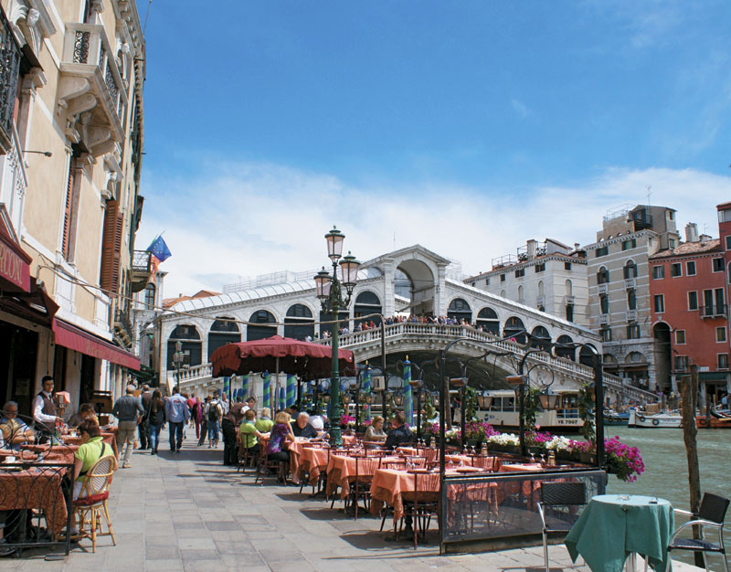 Restaurant-goers in Venice have lunch outdoors with views of canal boats cruising under the Rialto Bridge