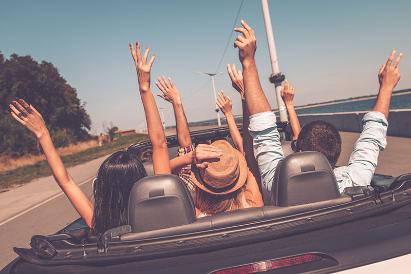 Rear view of young happy people enjoying road trip in their convertible and raising their arms up
