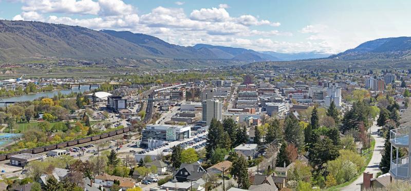 Panoramic view of downtown Kamloops with mid-rise buildings, a river, a freight train, and surrounding hills under a partly cloudy sky.