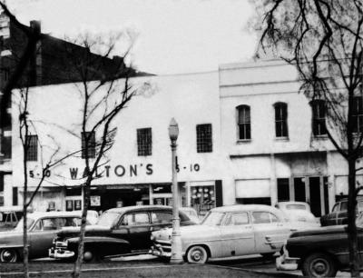 Black-and-white photo of a small shop on a midcentury American street. The shop sign above the windows, black letters on a white background, says "Walton's 5-10." Fantastic midcentury sedans are parked front-end-in on the street in front.