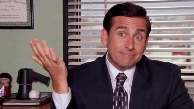 Michael Scott from "The Office." Head-and-shoulders photo of a neatly dressed and groomed white man, wearing a neat suit and tie, befuddled expression on his face, right hand raised
