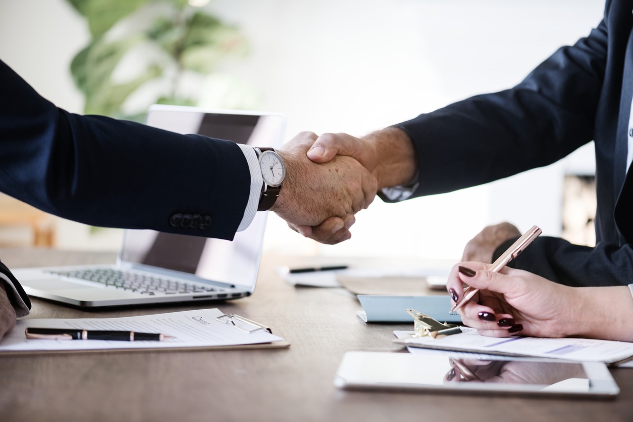 handshake over a desk with computers and someone taking notes